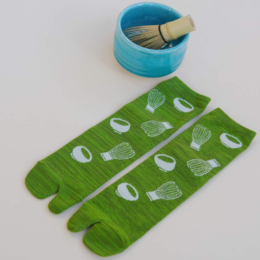 Green tabi socks with white patterns next to a blue bowl and brush on a light gray background