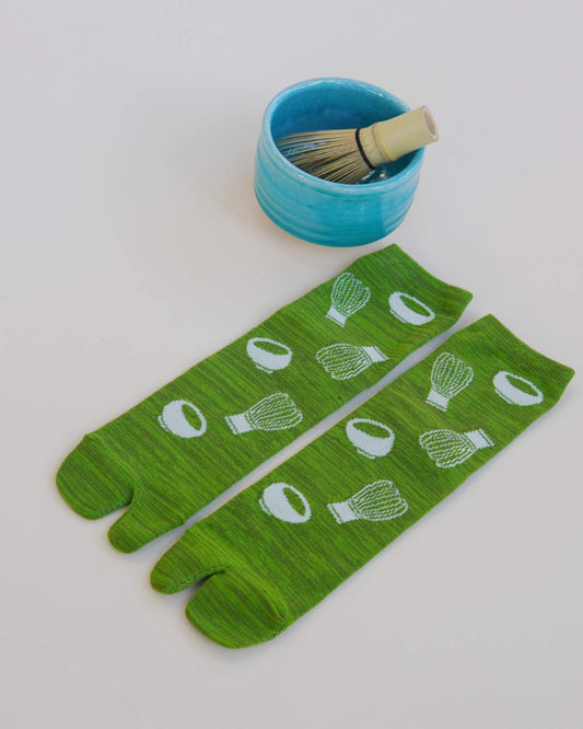 Green tabi socks with white patterns next to a blue bowl and brush on a light gray background